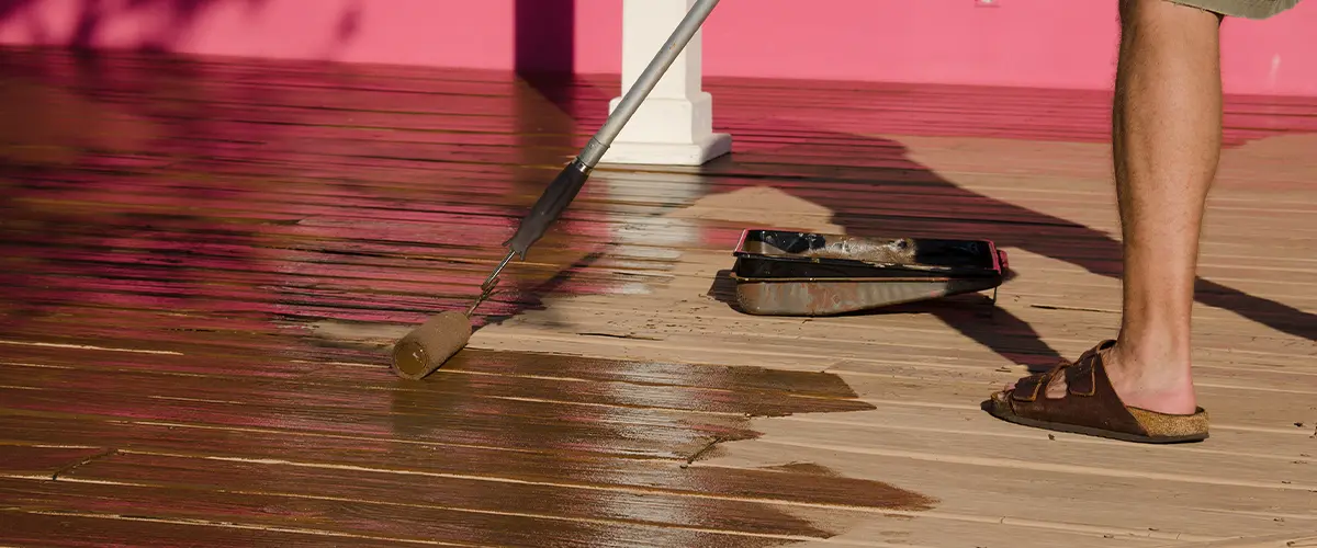 Man applying stain on wooden deck with roller illustrating Spraying Deck Stain Vs Rolling technique.