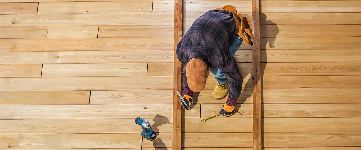 Carpenter measuring and marking wooden planks during deck installation, top view of worker with tools and materials.