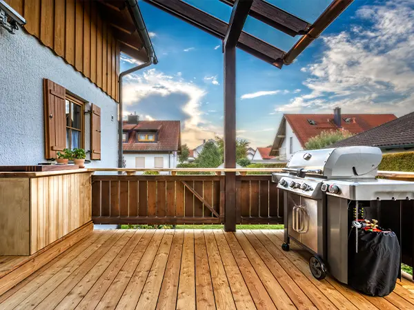 Charming wooden deck with a gas grill and planter boxes, overlooking a picturesque neighborhood under a clear blue sky.