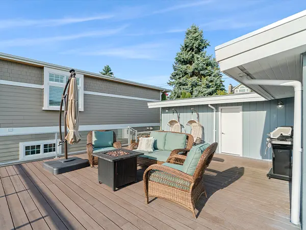 Modern backyard deck with wicker furniture and fire pit table, shaded by an umbrella, featuring cozy seating and a gas grill.