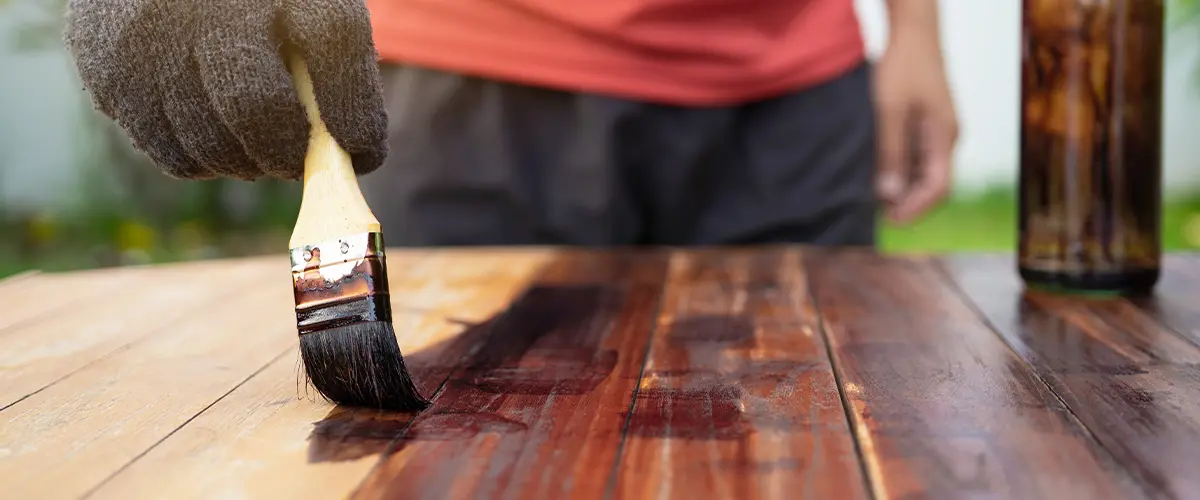 Person in a red shirt and gloves staining a wooden surface outdoors using a brush, emphasizing wood finishing and outdoor projects.