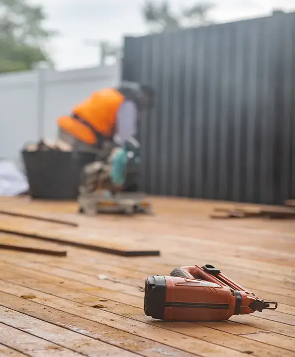 Close-up of a nail gun on a wooden deck under construction, with a worker in an orange vest using a saw in the background