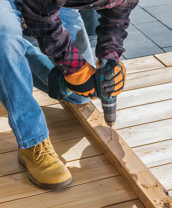 Worker in gloves and boots using a power drill to secure a wooden deck board during construction