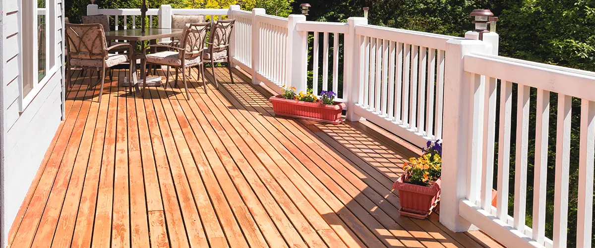 Sunny outdoor deck with white railings, a patio table set under an umbrella, and potted plants, surrounded by lush greenery