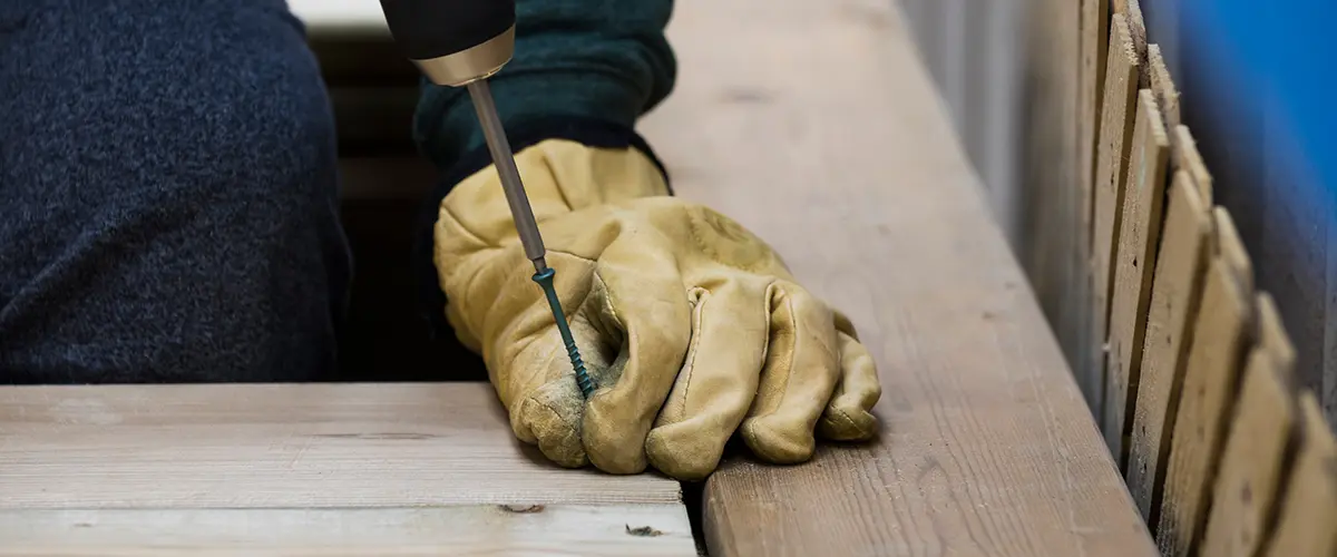 Close-up of a worker's hand wearing gloves while using a power drill to screw a deck board in place.