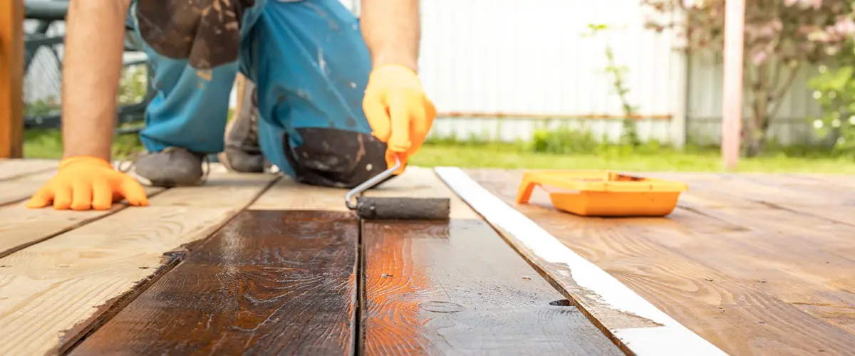 Man applying wood stain with roller to outdoor deck during renovation.