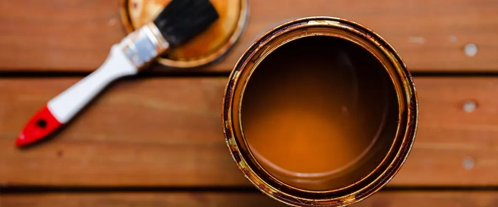 Open can of wood stain with a brush on a wooden deck, highlighting deck refinishing and maintenance process.