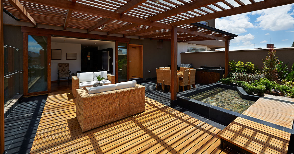 Modern wood patio with pergola, outdoor seating, dining area, and water feature surrounded by lush landscaping